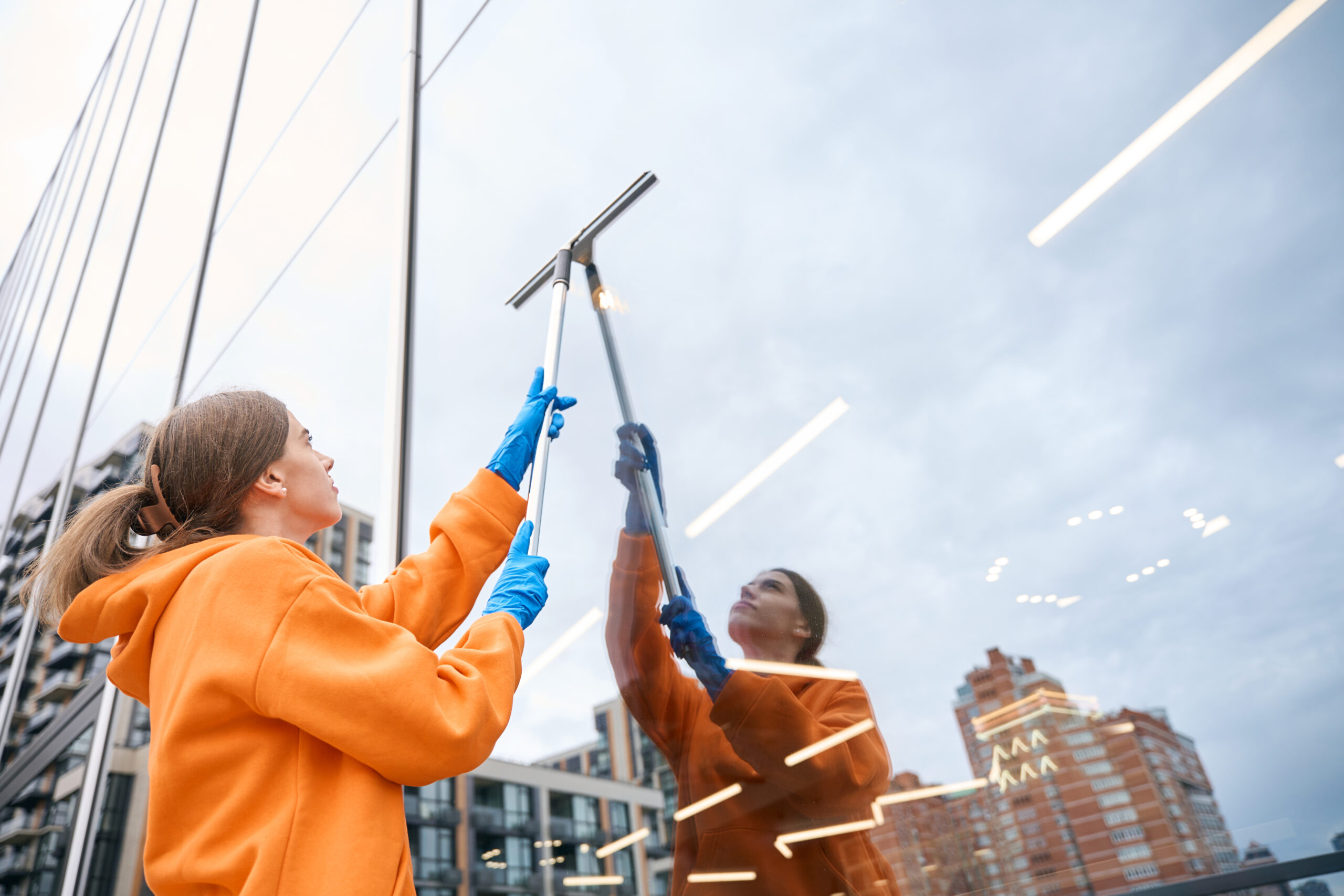 Young cleaning lady in a yellow hoodie and blue gloves cleans large windows with a long-handled window cleaning mop